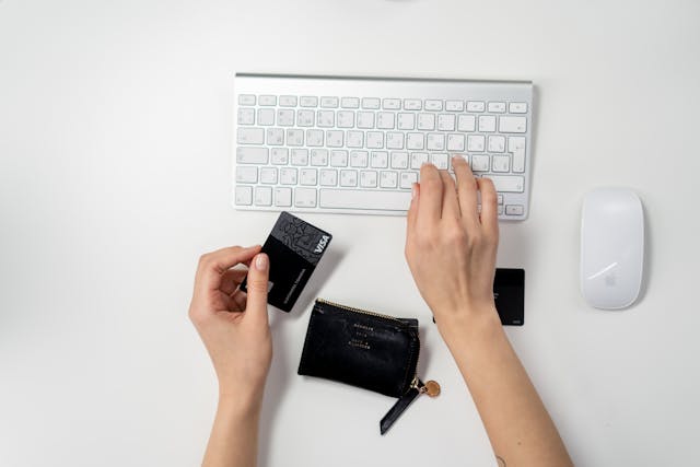 A person holding a credit card in one hand and typing on a computer keyboard with the other to buy something online.